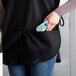 A woman holding a device in the pocket of a black Choice cobbler apron.