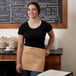 A woman wearing a tan Choice standard waist apron with 3 pockets stands in front of a chalkboard menu.