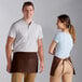 A man and woman standing next to each other wearing brown Choice standard waist aprons with 3 pockets.