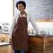A smiling woman wearing a Choice brown poly-cotton bib apron standing in front of a chalkboard.