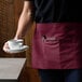 A woman wearing a burgundy standard waist apron with three pockets holding a cup and saucer.