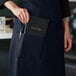 A woman wearing a navy blue apron with a notepad in the pocket.