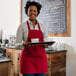 A black woman wearing a red Choice Poly-Cotton bib apron holding a tray of coffee cups.