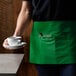 A person wearing a Choice Kelly Green standard waist apron holding a cup of coffee.