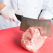 A hand in a glove uses a 9" stainless steel meat cleaver to cut a piece of meat on a cutting board.