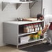 A man in a professional kitchen using a stainless steel table with a shelf full of food.