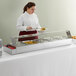 A woman in a white apron using an Avantco glass drop-in food warmer to serve food on a buffet.