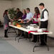 A group of people serving food on a Lifetime almond plastic folding table.