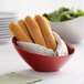 A red slanted melamine bowl filled with bread sticks on a table with a salad.