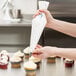 A person holding a pastry bag decorates cupcakes with white frosting.