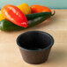 A black HS Inc. polyethylene ramekin on a wood surface containing food next to a pile of red peppers.