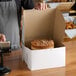 A person holding a 10" x 10" white cake box on a bakery counter.
