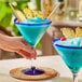 A hand holding a blue cocktail in an Acopa Tropic martini glass with a blue stem and rim.