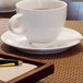 A white cup of brown liquid on a white saucer on a table with a pen and notepad.