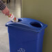 A person putting a bottle into a blue Carlisle recycling bin with a rectangular lid.