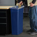 A person standing next to a Carlisle blue rectangular trash can.