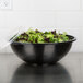 A Sabert black round bowl filled with lettuce with a plastic lid.