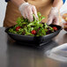 A person in gloves using a Fineline black plastic bowl to serve salad.