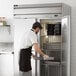 A man in a black apron and uniform opening a Beverage-Air reach-in refrigerator with a white background.