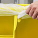 A hand holding a yellow plastic bag over a Rubbermaid yellow round trash can.
