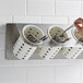 A hand holding a spoon in a Steril-Sil stainless steel flatware organizer with white perforated cylinders.