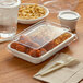 A rectangular plastic container with a white lid filled with food including chicken wings and french fries on a table.