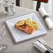 A Visions square white plastic plate with a piece of salmon and a lemon wedge on it, with a fork.