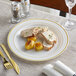 A Visions plastic plate with gold bands holding a piece of meat and a piece of bread with herbs on a table.