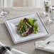 A Visions square white plastic plate with silver bands holding a salad on a table with silverware.