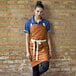 A woman wearing a Uncommon Chef Walnut Canvas Bib Apron with natural webbing and 3 pockets leaning against a brick wall.