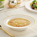 A Visions bone plastic bowl with gold bands filled with soup on a table with a spoon.