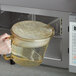 A hand holding a large glass container with liquid pouring into a microwave.