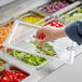 A hand using a Cambro clear plastic food pan lid with a spoon notch to cover a tray of food.