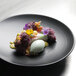 A Front of the House semi-matte black porcelain plate with dessert, flowers, and ice cream.