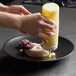 A person pouring yellow liquid onto a Front of the House Spiral Ink semi-matte black porcelain plate with a dessert.