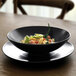 A Front of the House semi-matte black porcelain plate with a bowl of pasta on it.