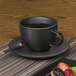 A black Front of the House porcelain cup with liquid in it on a table with a plate of fruit.