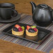 A black Front of the House rectangular porcelain plate with fruit tarts on it on a table with a black teapot.