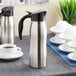A silver stainless steel insulated coffee carafe on a table with a cup of coffee.