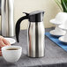 A hand pours coffee from a Choice stainless steel carafe into a white coffee cup.