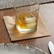 A glass with a drink and ice next to a piece of ice on a Hoffmaster Coin Embossed cocktail napkin.