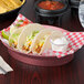 An oval raspberry deli server filled with tacos and a bowl of chips on a table with a red and white checkered napkin.