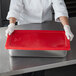 A person wearing a white coat and gloves holding a red Vollrath Super Pan V lid over a stainless steel container.