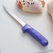 A Dexter-Russell purple narrow boning knife with a blue handle on a white surface next to a piece of chicken.