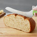 A gloved hand uses a Dexter-Russell green scalloped bread knife to cut a loaf of bread on a cutting board.