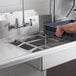 A person using a 20" x 20" slide bar to hold a blue container in a stainless steel sink on a counter.