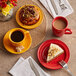 A table with a red plate of food and a mango orange Acopa Capri stoneware saucer with a cup of coffee on it.