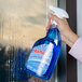 A hand holding a blue bottle of Noble Chemical Reflect Ready-to-Use Glass / Multi-Surface Cleaner and spraying a window.