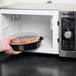 A hand placing a Pactiv Newspring VERSAtainer oval plastic container of food in a microwave.