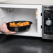 A hand placing a black Pactiv Newspring VERSAtainer oval plastic container of food in a microwave.